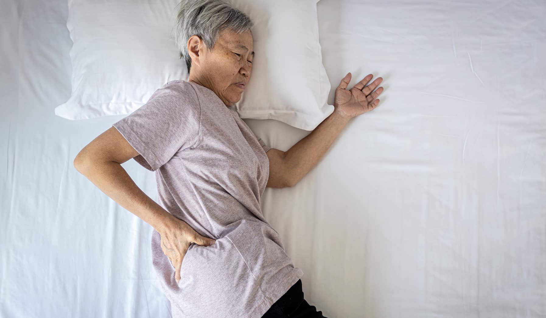 an elderly woman with lower back pain is resting her head on a bed with a pillow and her hand massaging her lower back