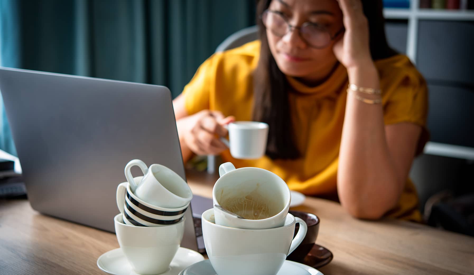 a stack of 5 differently sized coffee and espresso cups sits in the foreground as a distressed woman frowns at her computer screen with a cup of coffee held in her hand