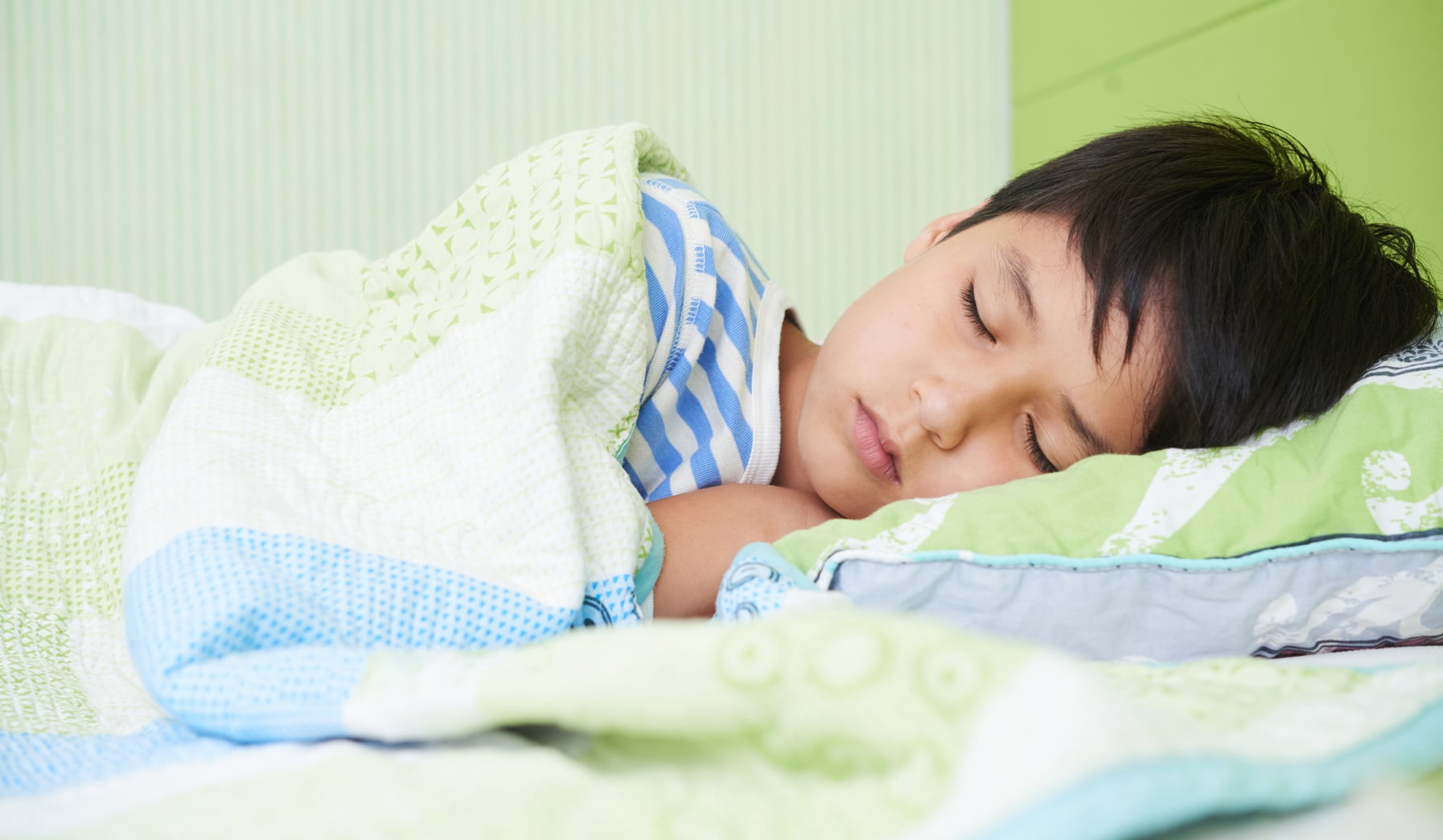 a small boy with black hair rests peacefully on a quilted pillow and under a quilted bed comforter that is in soft pastel colors of green and blue