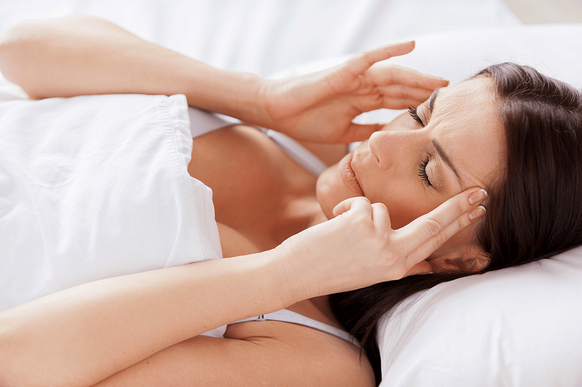 woman in bed is massaging each side of her head with her fingers as her head rests on a pillow with eyes closed