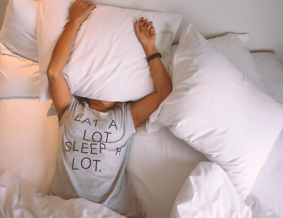 a woman sleeping on a bed with a pillow over her face. She's wearing a tshirt that says "eat a lot. sleep a lot."