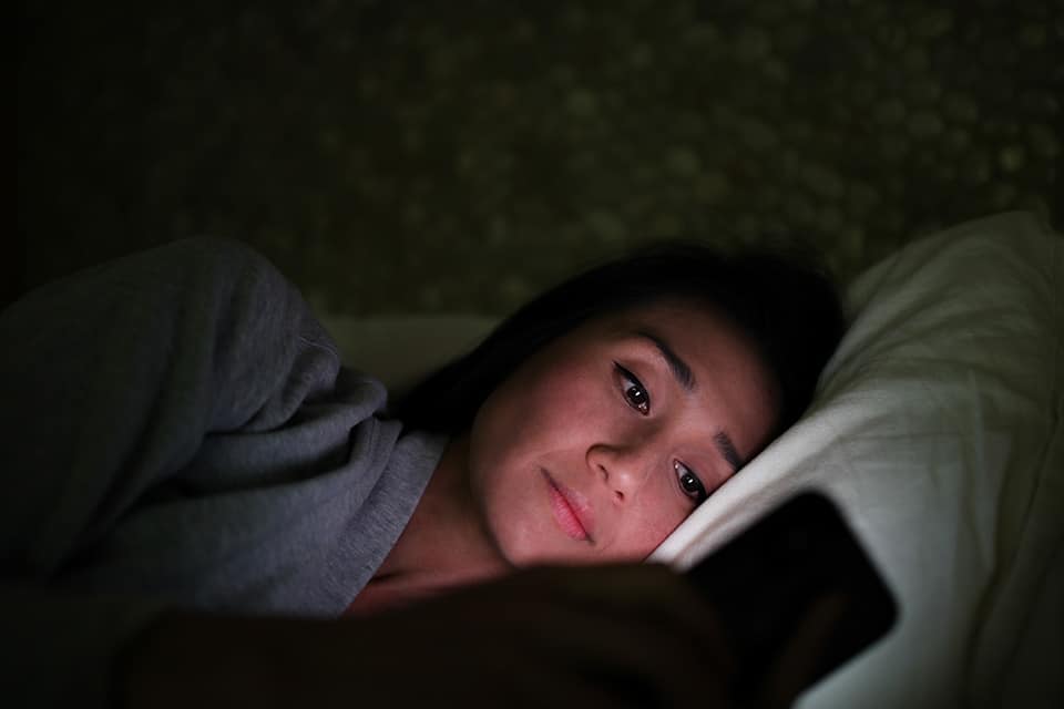 woman looking at her phone with her head against the pillow on her bed. The lights are all off with exception of the glow from her smart phone screen as she views the screen
