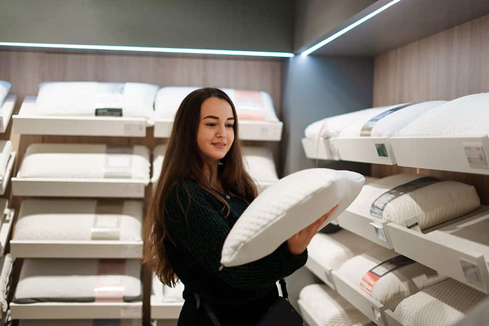 Cheerful brunette lady choosing new pillows for family, standing in bedding store department examines ergonomic design, adjustable memory foam, different fillings and fabrics for better quality sleep